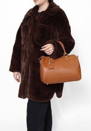 Manteau en fausse fourrure marron avec de grandes poches avant, porté avec un sac à main en cuir beige texturé comportant une fermeture éclair et des étiquettes décoratives.