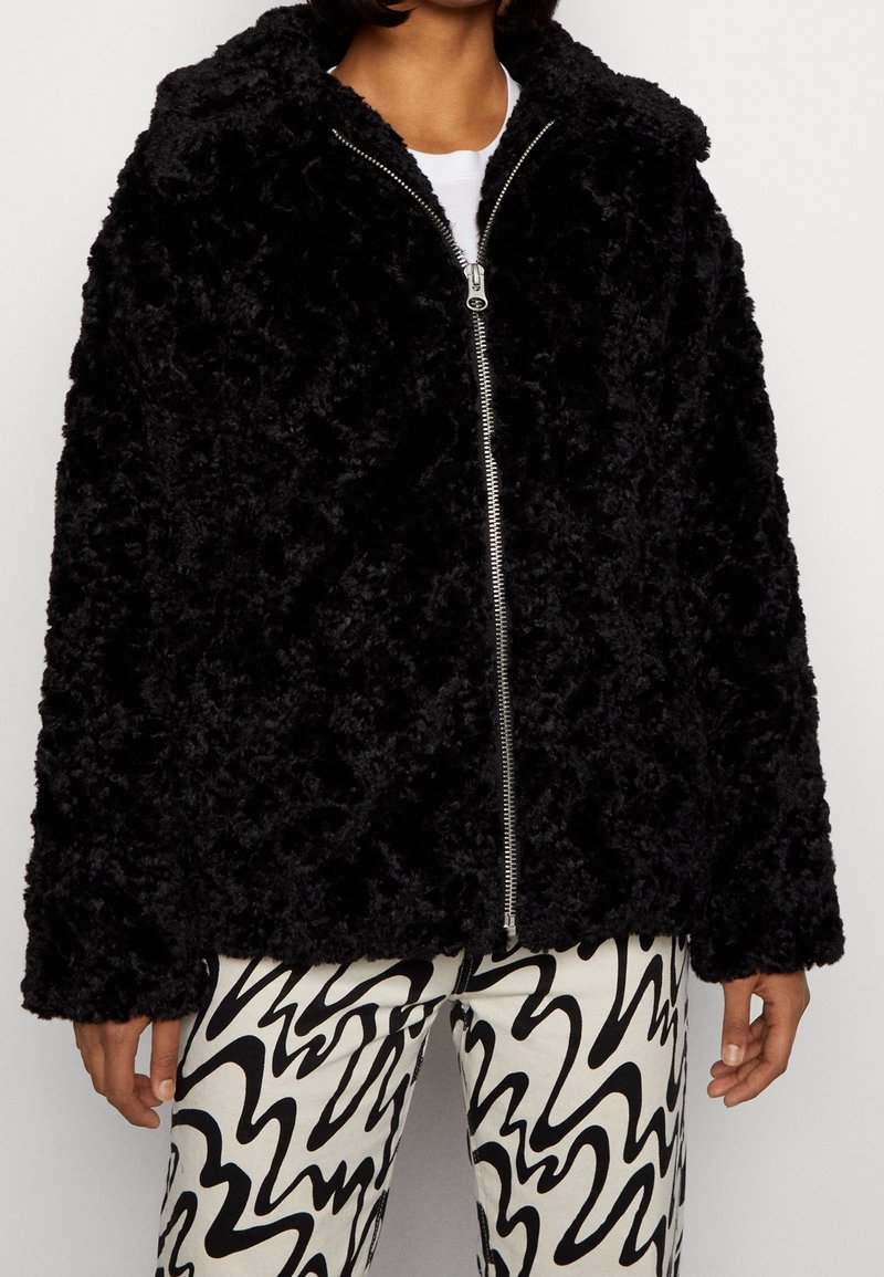 Personne portant une veste noire duveteuse zippée par-dessus une chemise blanche et un pantalon aux motifs abstraits noir et blanc, debout devant un fond uni.
