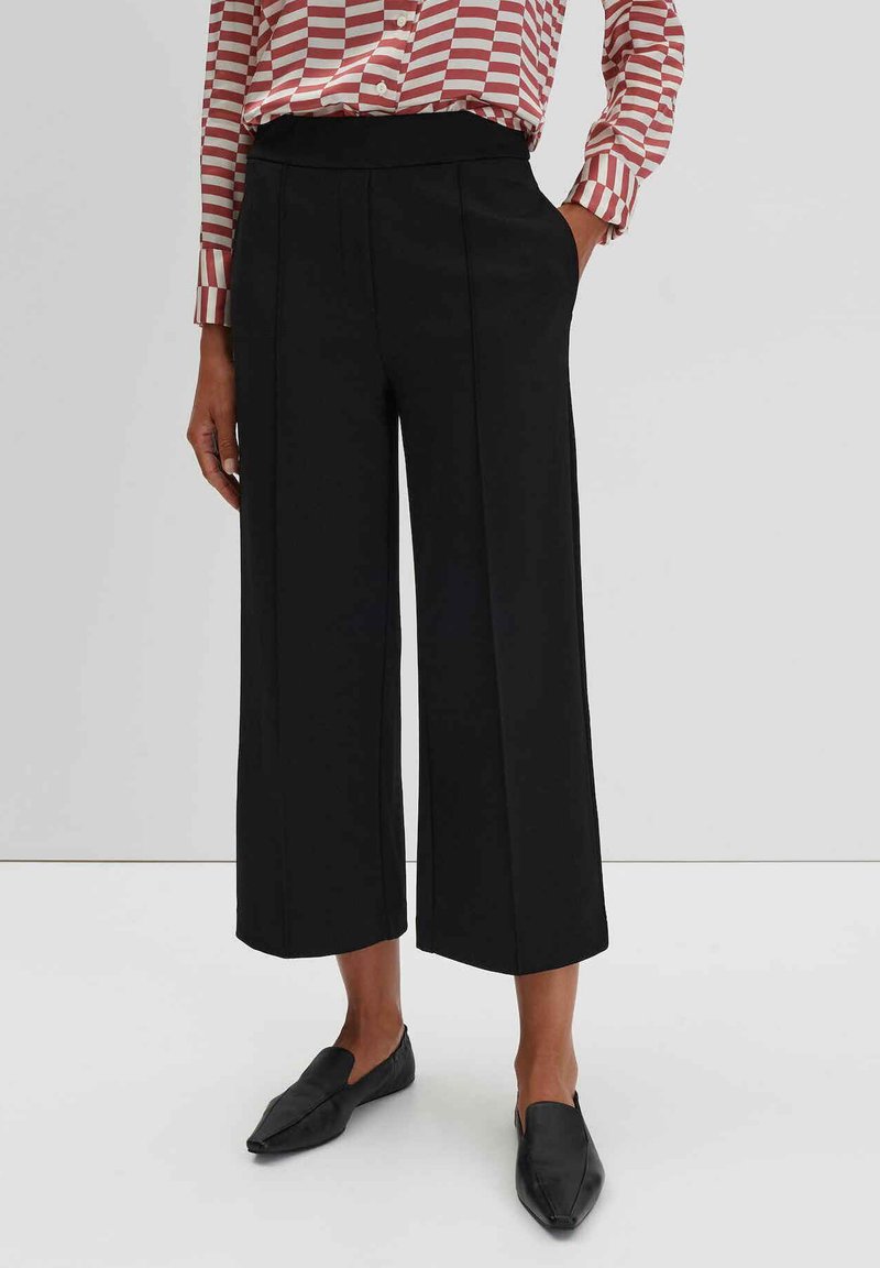 Zwarte wijde broek met een gladde textuur, voorzien van voorplooien en een elastische tailleband, gecombineerd met vlakke, puntige zwarte schoenen.