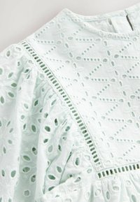 Tissu vert menthe clair avec des motifs floraux brodés, présentant des bords festonnés et des découpes délicates, mettant en valeur la texture et les détails.