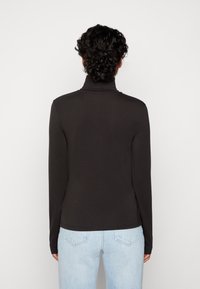 Person med lockigt mörkt hår som bär en svart långärmad polotröja och ljusblå jeans, vänd bort från kameran mot en enfärgad vit bakgrund.