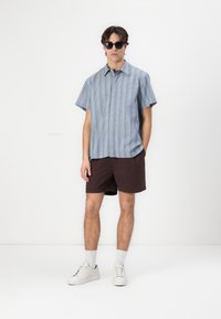 Kurze Ärmel, kragenloses Hemd in Blau mit vertikalen Streifen, kombiniert mit dunkelbraunen Shorts. Das Model trägt Sonnenbrillen und weiße Sneaker.