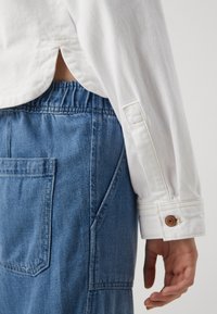 Vit långärmad skjorta med manschetter, stoppad i blå denimoveraller med en stor ficka och elastisk midja.