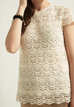Beige kanten top met een gekartelde zoom, met ingewikkelde golfpatronen en korte mouwen. Zachte, gestructureerde afwerking met een ronde hals.