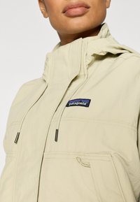 Ελαφριά μπεζ μπουφάν Patagonia με ψηλό γιακά, δύο μπροστινές τσέπες και διακριτικό λογότυπο. Διαθέτει ρυθμιζόμενα κορδόνια και απαλή υφή.