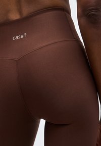 Καφέ αθλητικό κολάν με ψηλή μέση που φοριέται από άτομο, με το λογότυπο "casall" στη μέση.