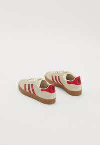 Sapatilhas de camurça com parte superior bege, riscas vermelhas e roxas, e um detalhe de calcanhar vermelho. Sola de borracha. Inclui cordões e detalhe do logótipo bordado.