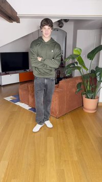 Vihreä teksturoitu huppari, harmaat farkut ja valkoiset tennarit. Taustalla ruskea corduroy-sohva ja suuri lehtikasvi. Puulattia.