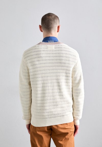 Helle beige gestrickte Pullover mit lockerer, strukturiertem Muster, gerippten Bündchen und Rundhalsausschnitt, über einem blauen Denimhemd getragen.