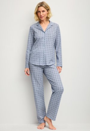 Femme blonde debout pieds nus portant un haut de pyjama boutonné à manches longues à motif bleu clair et un pantalon assorti sur un fond uni.