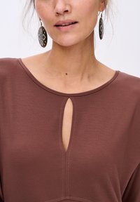 Haut marron avec un col rond, présentant un petit détail en forme de clé. Texture lisse avec des accents de couture subtils, associé à des boucles d'oreilles à motifs.
