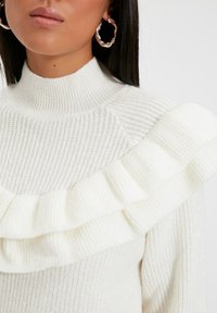 Weißer Strickpullover mit hohem Kragen, strukturiertem Körper und einem auffälligen Rüschendetail an der Schulter. Runde silberne Ohrringe.