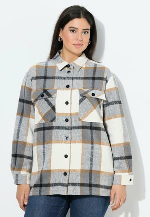 Giovane donna con lunghi capelli scuri che indossa orecchini a cerchio in argento e una camicia a quadri beige, grigia e nera sopra dei jeans blu.