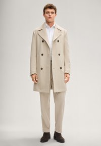 Beiger doppelt geknöpfter Mantel aus strukturiertem Stoff, mit großen braunen Knöpfen, breiten Revers und geradem Schnitt, kombiniert mit hellfarbigen Hosen.