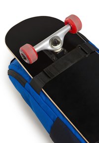 Skateboard con tavola nera, truck argentati e ruote rosse, poggiato su uno zaino blu con spallacci neri. La texture è liscia e robusta.
