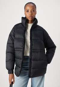 Casaco puffer preto com colarinho alto, fecho de correr e punhos elásticos. Apresenta seções acolchoadas e um corte relaxado. Usado sobre um suéter creme.