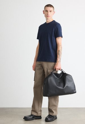 Schwarze synthetische Sporttasche mit abgerundetem Deckel, kurzen Griffen und sichtbaren Stickdetails, kombiniert mit einem navyblauen Rundhalsausschnitt-T-Shirt und beigen Cargo-Hosen.