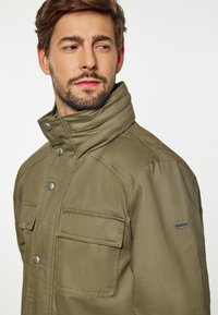 Olivengrøn jakke med høj krave, trykknapper og to forlommer. Glat stof med let glans. Har et logo-patch på ærmet.