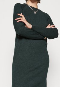 Abito maglione di colore verde scuro con collo a coste e maniche lunghe. Presenta spacchi laterali per una maggiore libertà di movimento e una texture morbida.