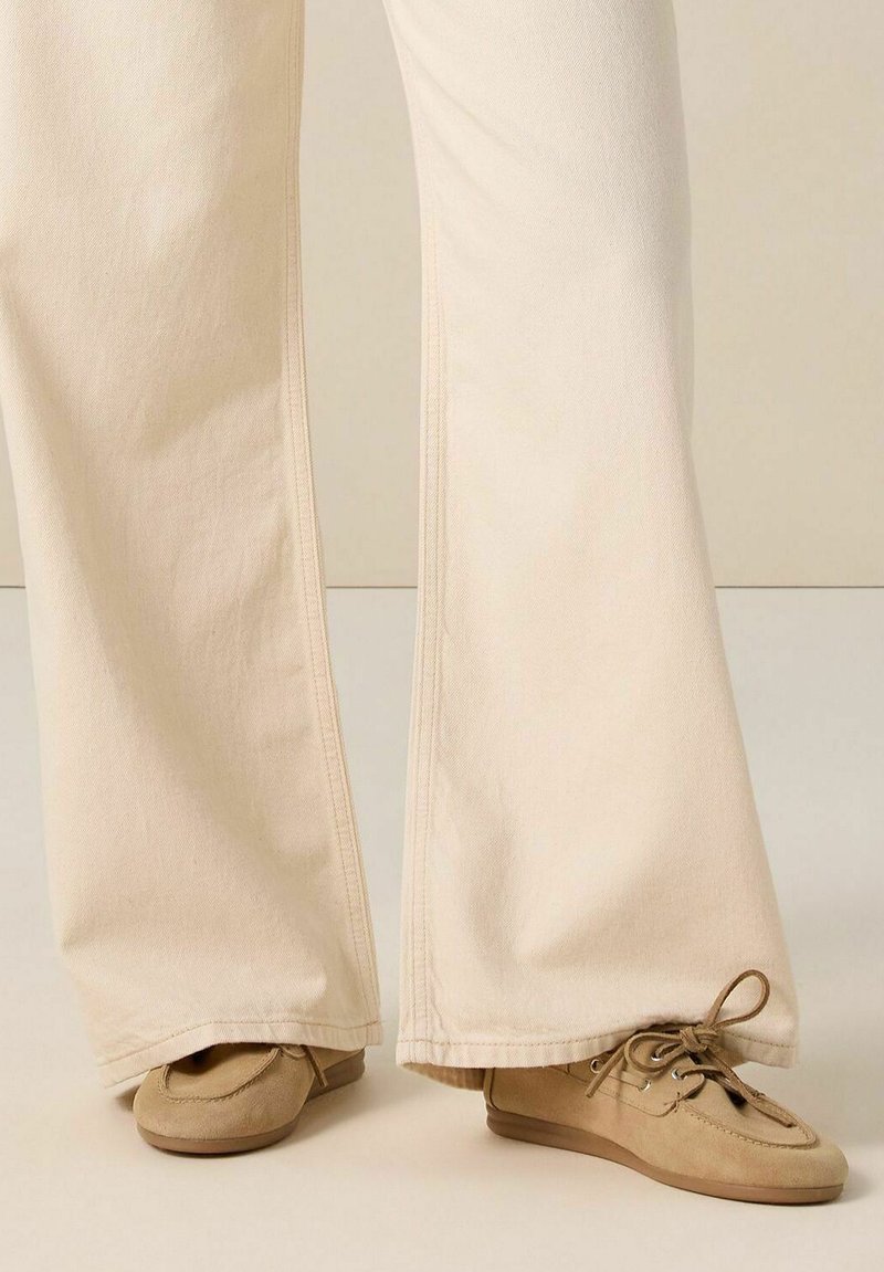 Pantalons beige à jambes larges avec une texture lisse, dotés de coutures contrastées, associés à des chaussures de style mocassin marron clair avec des lacets.