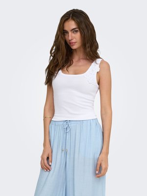 Femme aux longs cheveux bruns portant un haut blanc sans manches avec un détail en dentelle sur une épaule et un pantalon bleu clair à cordon.