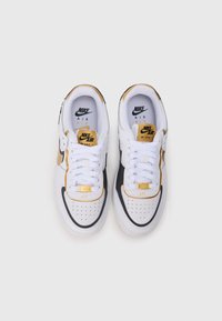 Para białych sneakersów Nike Air Force 1 z czarnymi i złotymi akcentami, białymi sznurowadłami oraz oznakowaniem na języku i sznurowadłach, widziana z góry.
