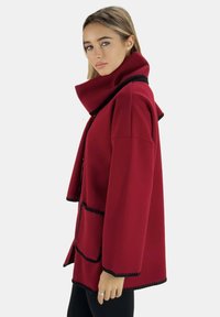 Abrigo rojo con cuello alto y ribete negro, con un bolsillo frontal. Fabricado con una tela texturizada, corte inclinado y un ajuste relajado.