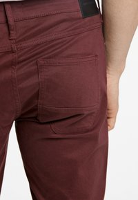 Bordeaux kleurige broek van gladde stof, met een achterzak, contrasterende stiksels en een leren label op de taille.