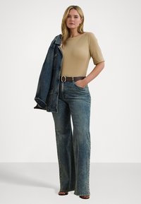Top bej ajustat, blugi evazați cu talie înaltă de culoare albastră, jachetă din denim purtată pe umăr, curea maro și sandale cu toc. Textura materialului este netedă.