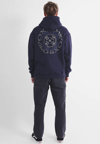 Sweat à capuche marine avec un grand design graphique au dos, comportant du texte et un motif circulaire. Associé à un pantalon sombre et des baskets noires.