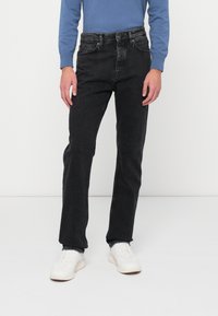 Črne jeans hlače s ravnim krojem, ki vključujejo pet �žepov in sprednji gumb, v kombinaciji z modro srajco z dolgimi rokavi in belimi športnimi copati.