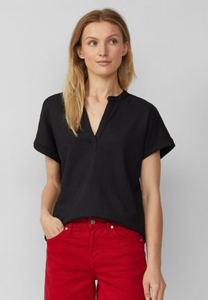 Schwarzes Kurzarmoberteil mit gerippter Textur, V-Ausschnitt und aufgerollten Ärmelbündchen, kombiniert mit roten, figurbetonten Hosen. Einfaches und lässiges Design.