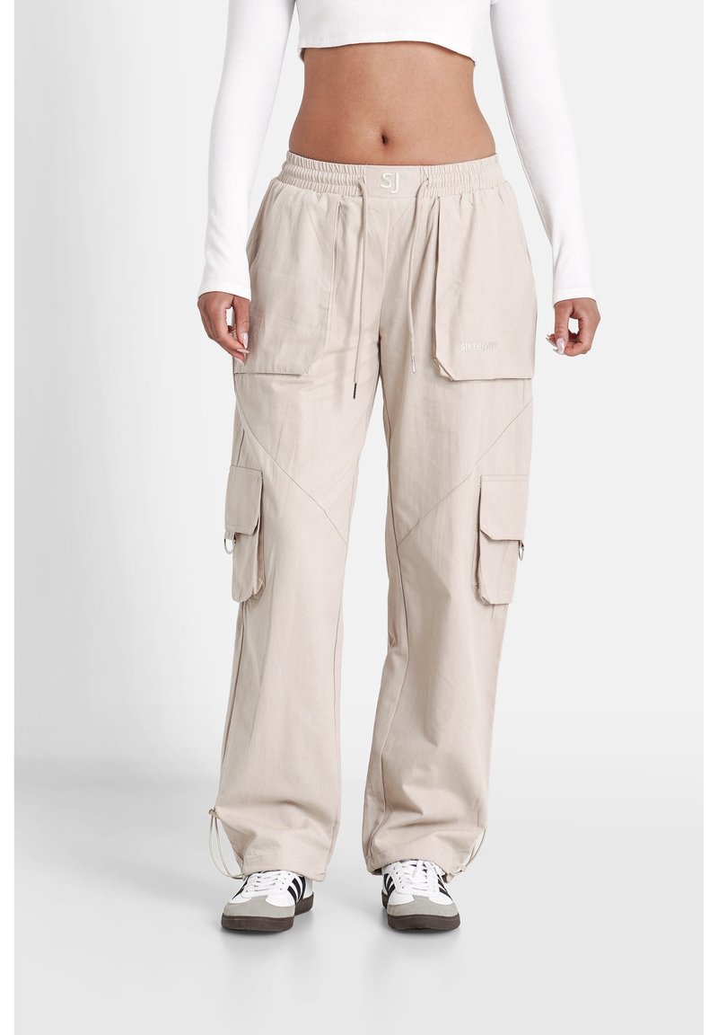 Femme portant un pantalon cargo beige avec plusieurs poches, taille à cordon et baskets blanches, montrée de la taille aux pieds.