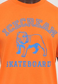 Oranžinė medvilninė marškinė su mėlynos spalvos grafika, kurioje pavaizduotas buldogas, ir įžymiu tekstu „ICECREAM SKATEBOARD D.“ Paprastas apvalaus kaklo dizainas.