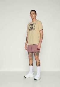 Beige cotton t-shirt with a black and camo print logo. Paired with mauve shorts and white sneakers, featuring a simple athletic design.