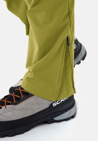 Pantalon de plein air vert avec une fermeture éclair latérale partiellement ouverte sur des chaussures de randonnée gris et noir avec des lacets orange sur fond blanc.