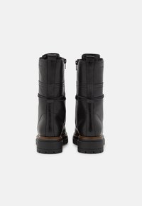 Par de botas de cuero negro hasta el tobillo con suelas gruesas y tiradores en la parte posterior, mostradas desde atrás sobre un fondo blanco.