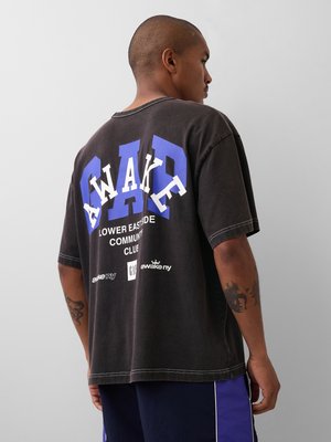 Hombre con tatuajes que lleva una camiseta negra de gran tamaño con la palabra "AWAKE" y otro texto en blanco y azul, de espaldas contra un fondo liso.