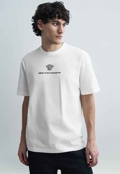 T-shirt en coton blanc avec col rond, manches courtes, présentant un graphisme noir de tête de Méduse et un logo texte centré sur le devant.