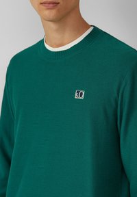 Sweter w kolorze niebieskozielonym z prążkowanym wzorem, z okrągłym dekoltem, białym wnętrzem kołnierza oraz małą haftowaną naszywką na piersi.