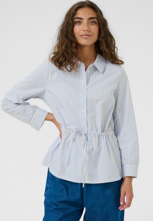 Žena s kučeravými hnedými vlasmi, oblečená v svetlomodrej a béžovej pruhovanej košeli na gombíky s opaskom v páse a modrých nohaviciach, pózujúca s rukou na boku.