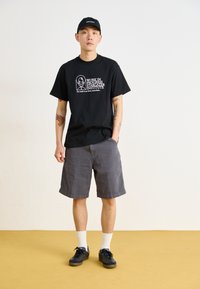 T-shirt preta com texto gráfico branco, calções cinzentos, meias brancas e ténis pretos. Roupa casual com um corte descontraído e tatuagens visíveis.