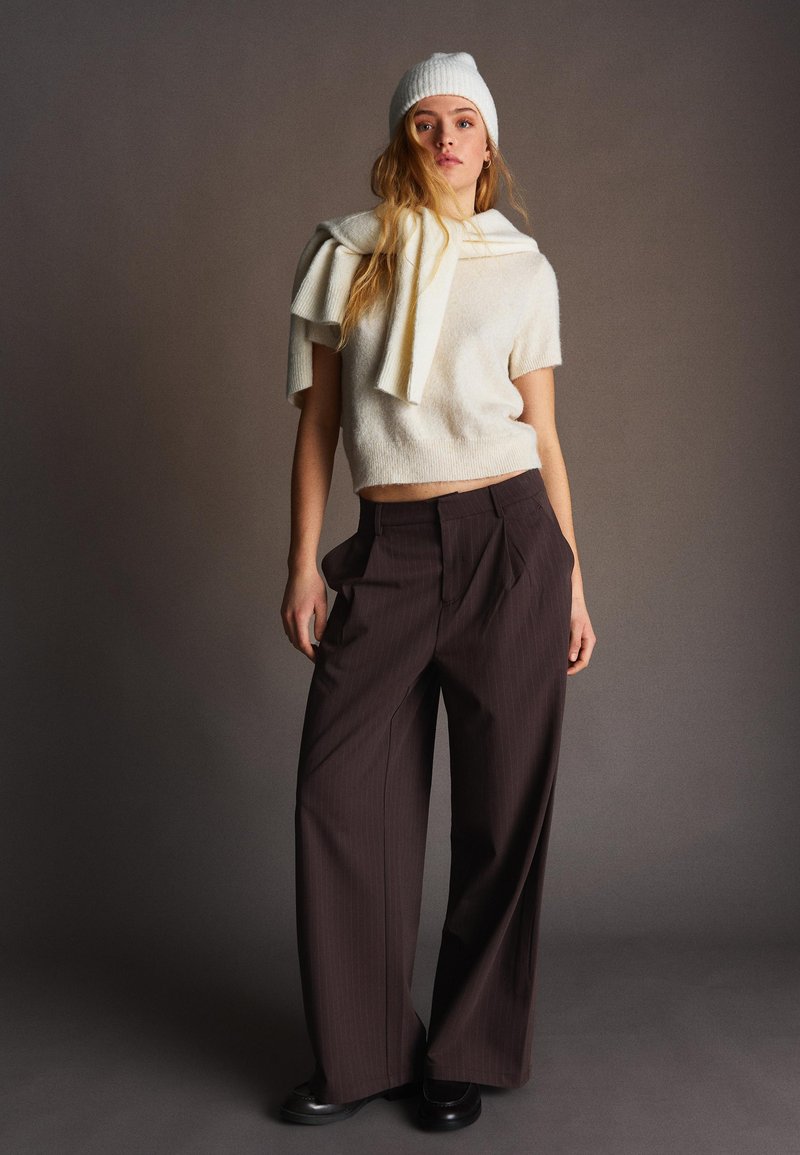 Jonge vrouw die een witte gebreide muts en trui draagt met een bijpassende trui over haar schouders, bruine broek met wijde pijpen en zwarte schoenen.