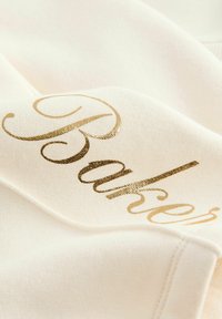 Bavlnená látka v krémovej farbe s lesklým zlatým nápisom "Baker." Mäkká štruktúra s viditeľným švom a záhybmi.