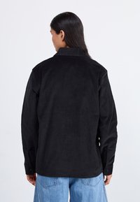 Camisa de pana negra con cuello, puños abotonados y un corte relajado, que presenta una superficie texturizada y un dobladillo recto.