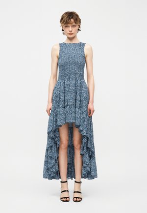 Kvinne iført en ermeløs blå blomstermønstret høyt-lavt kjole med volanger, kombinert med svarte høyhælte sandaler med stropper, stående mot en ensfarget bakgrunn.