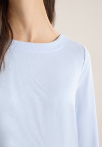 Femme portant un haut à manches longues côtelé bleu clair avec encolure ronde, montrant les détails du cou et des épaules sur un fond beige.