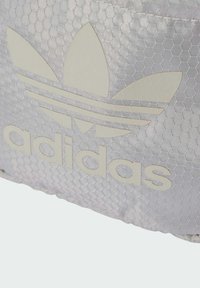Un tessuto argento con un motivo esagonale presenta un prominente logo trefoil Adidas e la parola "adidas" in tipografia beige chiaro.