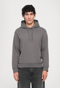 Jaunas vyras su garbanotais tamsiais plaukais, vilkintis pilką Armani Exchange džemperį su kapuche ir juodas kelnes, stovintis prieš paprastą baltą foną.