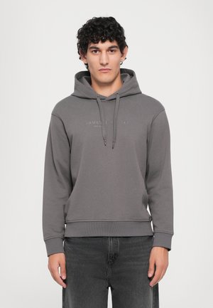 Junger Mann mit lockigem, dunklem Haar, der einen grauen Armani Exchange Hoodie und schwarze Jeans trägt, vor einem schlichten weißen Hintergrund stehend.